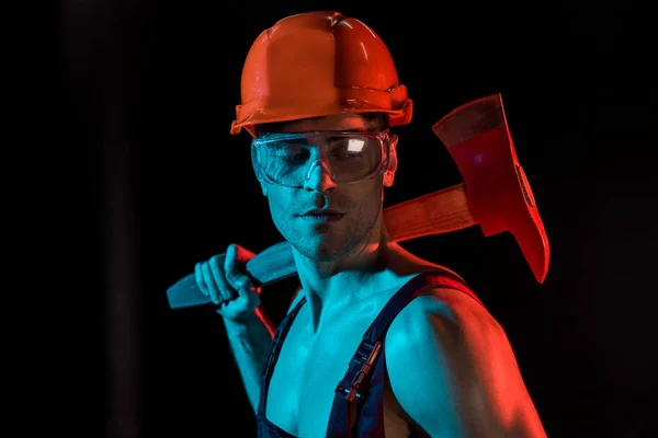 Shirtless fireman in overall, hardhat and protective goggles holding flat head axe on black — Stock Photo
