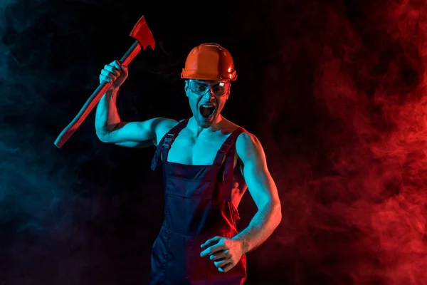 Besorgter Feuerwehrmann in Harthut und Schutzbrille, der eine flache Axt in der Hand hält und in Rauch auf schwarz schreit — Stockfoto