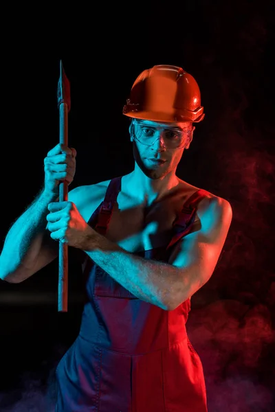 Serious shirtless fireman in hardhat and protective goggles holding flat head axe in smoke on black — Stock Photo