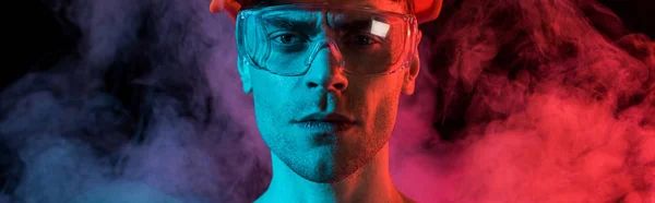 Panoramic shot of serious fireman in hardhat and protective goggles in red smoke on black — Stock Photo