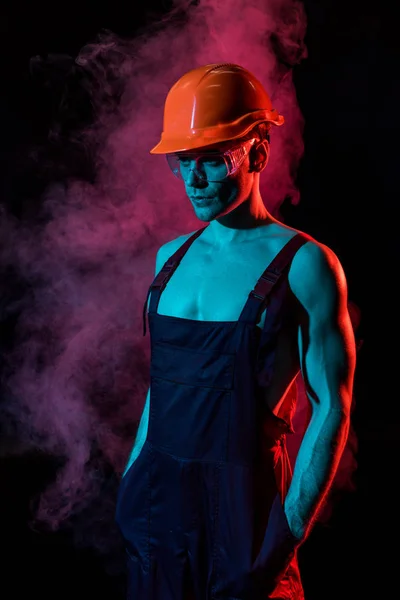 Sexy shirtless fireman in overall and hardhat with hands in pockets in smoke on black — Stock Photo