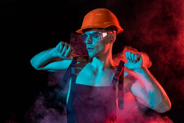 Sexy shirtless fireman in overall and hardhat in smoke on black — Stock Photo