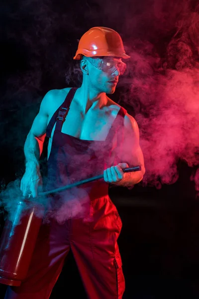 Sexy hemdloser Feuerwehrmann im Overall hält Feuerlöscher in Rauch auf schwarz — Stockfoto