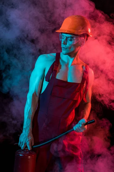Sexy shirtless fireman in overall holding fire extinguisher in smoke on black — Stock Photo