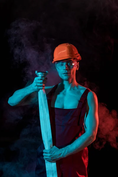 Sexy fireman in hardhat and protective goggles holding fire hose in smoke on black — Stock Photo