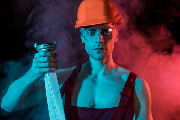 Sexy fireman in hardhat and protective goggles holding fire hose in smoke on black — Stock Photo