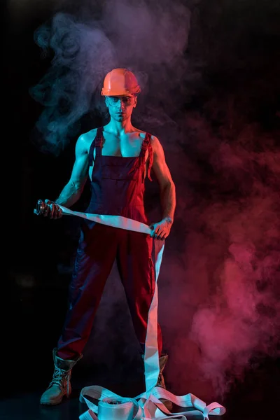 Full length view of sexy shirtless fireman holding fire hose in smoke on black — Stock Photo