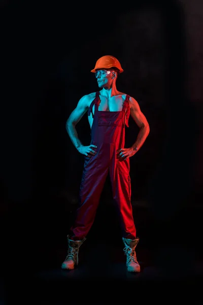 Full length view of sexy fireman in overall, hardhat and protective goggles with arms akimbo on black — Stock Photo