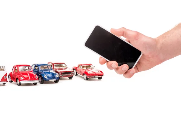 Vista recortada del hombre sosteniendo el teléfono inteligente con pantalla en blanco y coloridos coches de juguete en blanco - foto de stock