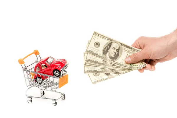 Vista cortada do homem segurando notas de dólar perto de carro de brinquedo vermelho no carrinho de compras isolado no branco — Fotografia de Stock
