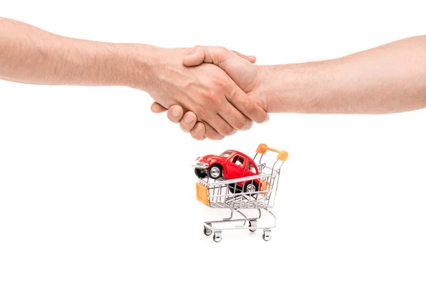 Vista recortada del hombre estrechando las manos cerca de coche de juguete en el carro de la compra aislado en blanco - foto de stock