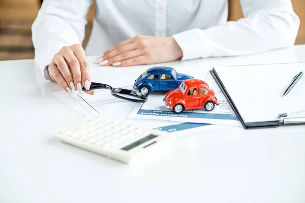 Cropped view of woman in formal wear holding magnifier at table with calculator, toy cars, clipboard and documents — Stock Photo