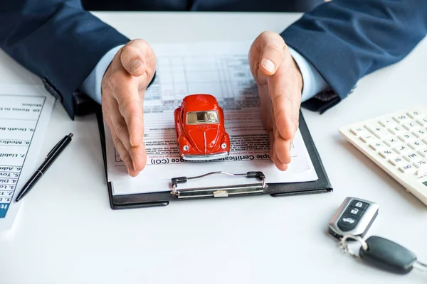 Vista recortada del hombre en ropa formal con coche de juguete rojo en la mesa con documentos, portapapeles, calculadora y llaves - foto de stock