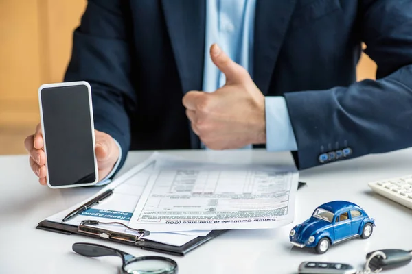 Partial view of man in formal wear holding smartphone with blank screen and showing thumb up at table with documents, blue toy car, clipboard, keys and magnifier — Stock Photo