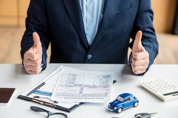 Vista recortada del hombre en ropa formal que muestra los pulgares hacia arriba en el lugar de trabajo - foto de stock