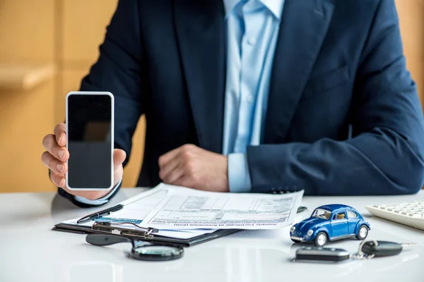 Vista parcial del hombre en desgaste formal celebración de teléfono inteligente con pantalla en blanco en la mesa con documentos, coche de juguete azul, llaves, portapapeles y lupa - foto de stock