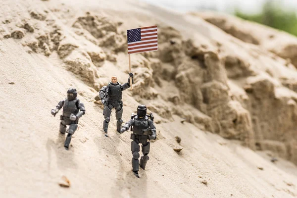 Toy soldiers with weapon standing on sand and holding american flag — Stock Photo