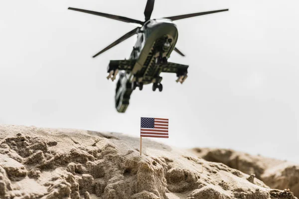 Enfoque selectivo de la pequeña bandera americana en la duna de arena con helicóptero militar por encima - foto de stock