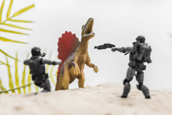 Selective focus of toy soldiers aiming guns on roaring tiny dinosaur on sand hill — Stock Photo