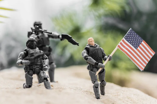 Selective focus of toy soldiers aiming with guns at toy man with american flag on sand hill — Stock Photo