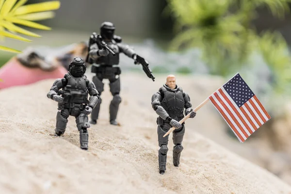 Selective focus of plastic toy soldiers aiming with guns at toy man with american flag on sand hill — Stock Photo