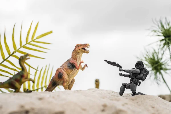 Foco seletivo do soldado de brinquedo plástico apontando com arma para dinossauro de brinquedo na duna de areia com folhas tropicais — Fotografia de Stock