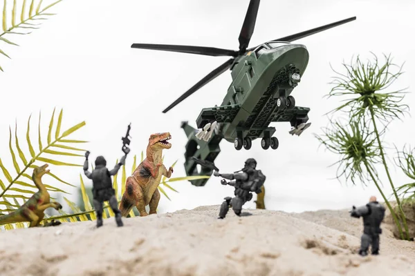 Selective focus of plastic soldiers aiming at toy dinosaurs on sand dune with helicopter in sky — Stock Photo