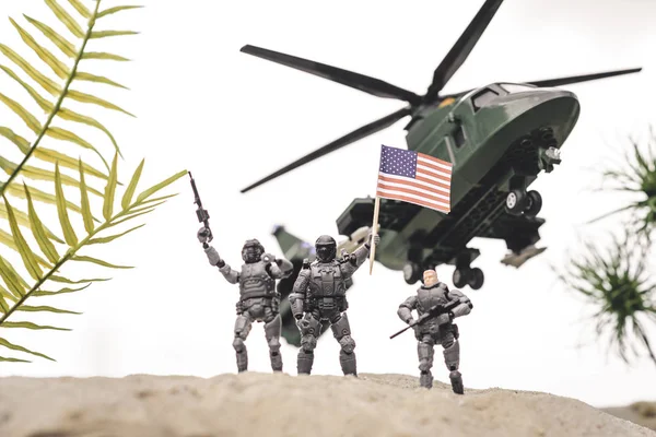 Soldados de brinquedo de plástico em duna de areia com armas e bandeira americana perto de helicóptero no céu — Fotografia de Stock