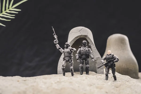 Soldats jouets avec des armes à feu et drapeau américain debout près des grottes sur une dune de sable sur fond noir — Photo de stock