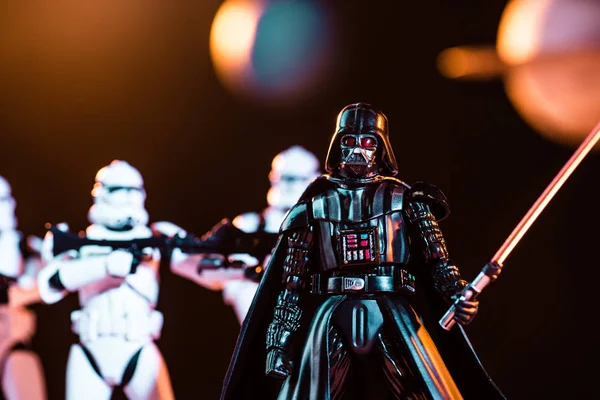 Selective focus of Darth Vader with lightsaber and white imperial stormtroopers with guns on background — стокове фото