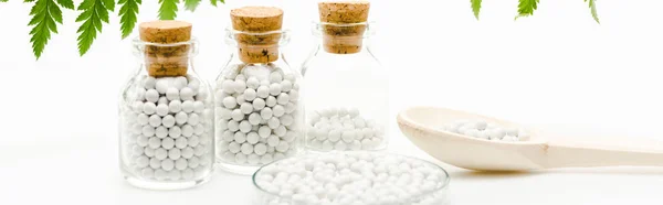 Panoramic shot of pills in glass bottles with wooden corks near green leaves on white — Stock Photo