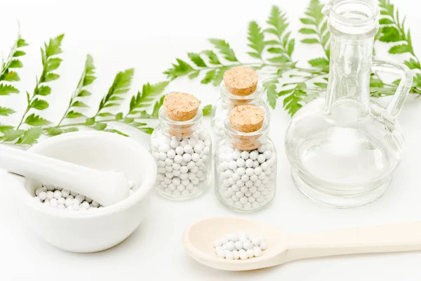 Bottiglie di vetro con piccole pillole vicino malta e pestello, cucchiaio di legno, vaso e foglie verdi su bianco — Foto stock
