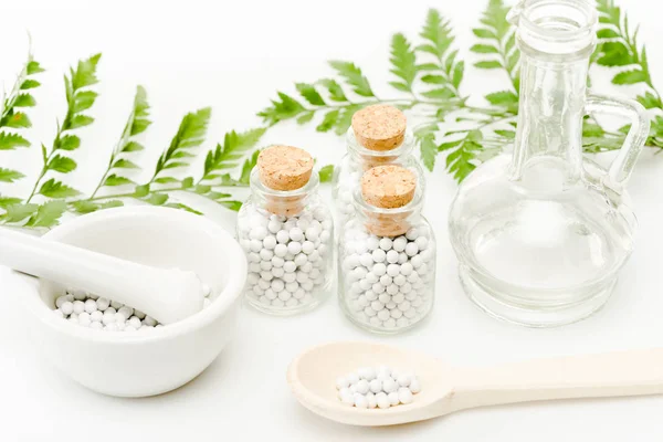 Frascos de vidrio con pequeñas píldoras cerca de mortero y pestle, cuchara de madera, frasco y hojas verdes en blanco - foto de stock