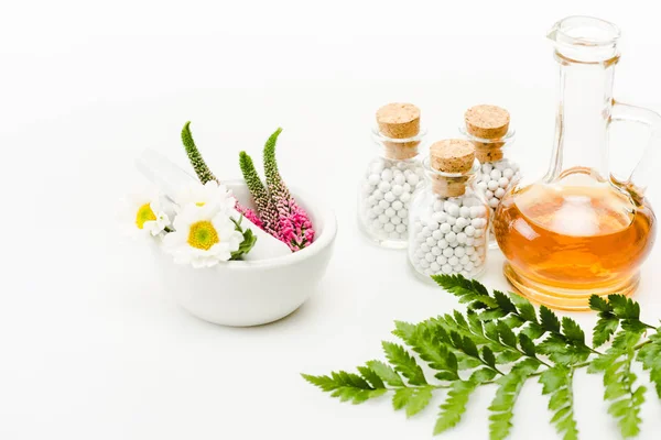 Flores em argamassa perto de pilão e garrafas de vidro com pílulas, jarro com óleo e folha verde sobre branco — Fotografia de Stock