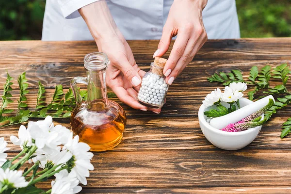 Vista cortada de mulher segurando garrafa com pílulas perto de mesa de madeira com plantas — Fotografia de Stock