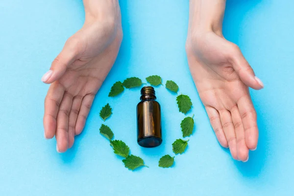 Cropped view of woman near glass bottle and leaves on blue — Stock Photo