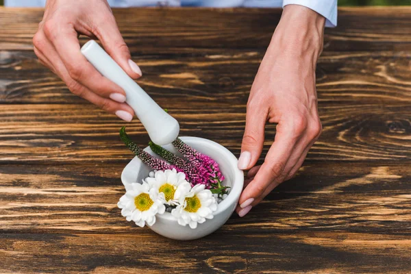 Cropped view of woman holding pestle near mortar with flowers on wooden table — Stock Photo