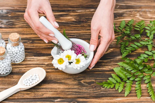 Cropped view of woman holding pestle near mortar with flowers and green leaves on wooden table — Stock Photo