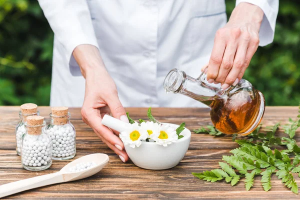 Vue recadrée d'une femme tenant une cruche avec de l'huile près du mortier avec des fleurs et des feuilles vertes sur une table en bois — Photo de stock