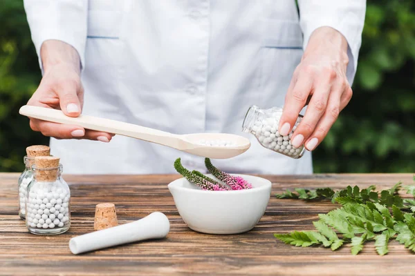 Cropped view of woman holding spoon and bottle with pills near mortar with veronica flowers and green leaves on wooden table — Stock Photo