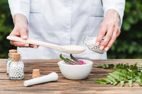 Vista recortada de la mujer sosteniendo cuchara y botella con pastillas cerca de mortero con flores de verónica y hojas verdes en la mesa de madera - foto de stock