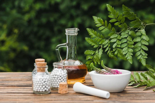 Messa a fuoco selettiva di pestello vicino malta con fiori, bottiglie di vetro con pillole e brocca con olio su tavolo di legno — Foto stock