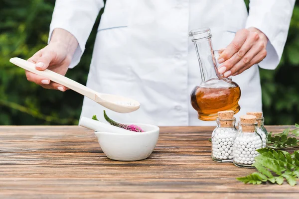 Vue recadrée d'une femme tenant une cuillère avec des pilules et une bouteille d'huile près du mortier avec des fleurs de veronica et des feuilles vertes sur une table en bois — Photo de stock