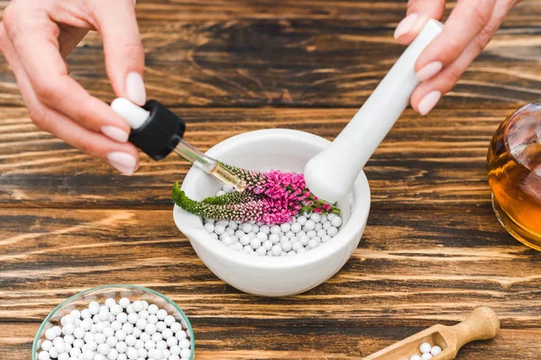 Vista recortada de la mujer sosteniendo pestle y pipeta cerca de mortero con flores verónica y pastillas en la mesa de madera - foto de stock