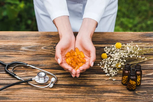 Cropped view of woman pills near bottles flowers and stethoscope on wooden table — Stock Photo
