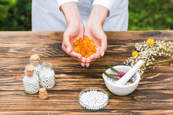 Vista recortada de la mujer sosteniendo píldoras cerca de botellas y mortero con flores de verónica en la mesa de madera - foto de stock