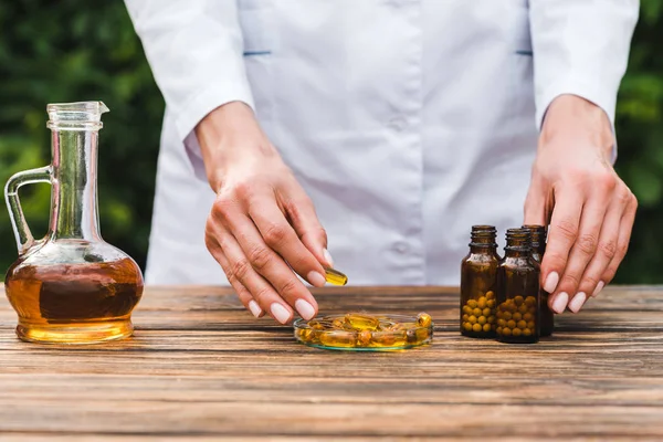 Cropped view of woman holding orange pill near bottles and jug with oil — Stock Photo