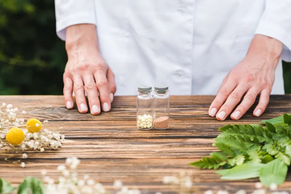 Cropped view of woman standing near wooden table with glass bottles near green leaves — Stock Photo