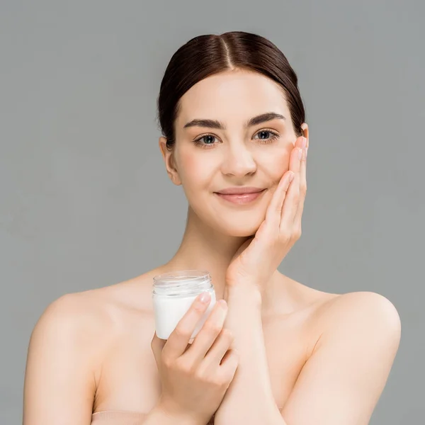 Cheerful naked woman holding container with cosmetic cream and touching face isolated on grey — Stock Photo