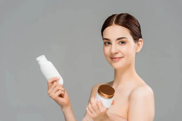 Cheerful brunette woman holding container with face cream and bottle with body lotion isolated on grey — Stock Photo